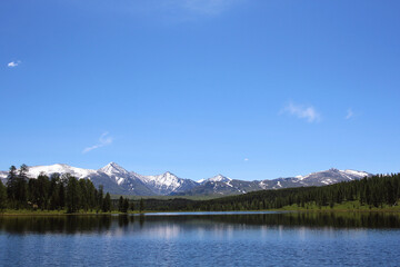 Fototapeta na wymiar The snow-covered mountain range and green forest are reflected in the crystal blue waters of the lake. Pastoral meditative landscape. Mountain Lake. Wildlife. Travel across Russia