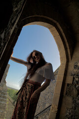A young lady with long brunette hair and hippie clothes poses in a castle archway