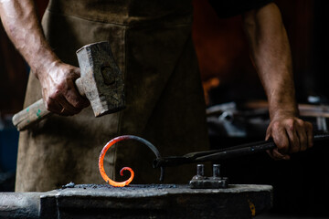 Blacksmith is processing a hot metal object of a spiral shape on the anvil in a dark forge