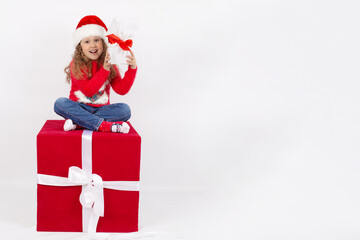 A little girl in a Santa Claus hat sitting on a red cube in the Studio with a gift in her hands, on a white background. The concept of a happy childhood. Copy space. Mock up