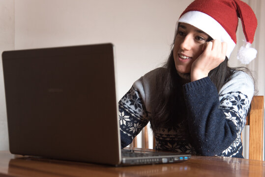 Una chica joven sonríe mientras trabaja con su ordenador portátil mientras lleva puesto un gorro de Santa Claus y un jersey de Navidad - Powered by Adobe