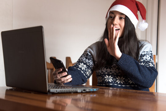 Una chica joven haciendo una video llamada saluda a su ordenador portátil mientras lleva puesto un gorro de Santa Claus y sujeta su teléfono móvil - Powered by Adobe