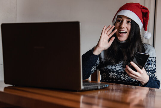 Una chica joven haciendo una video llamada saluda a su ordenador portátil mientras lleva puesto un gorro de Santa Claus y sujeta su teléfono móvil - Powered by Adobe