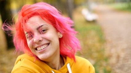 Portrait of a young pretty smiling woman with bright pink hair. Walk in the autumn park. Hipster attractive adult girl in oversized yellow hoodie or hoody. Positive emotions. Smile. 
