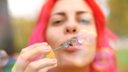 Happy adult girl blowing soap bubbles front camera in nature. Close-up. Good-looking girl blows bubbles at camera and has fun. Portrait young cheerful caucasian woman with bright pink hair. 