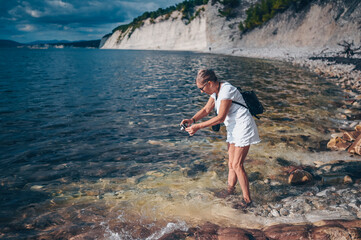 Elderly senior traveling backpacker mature woman tourist taking photos sea coast, rocks, blue sky