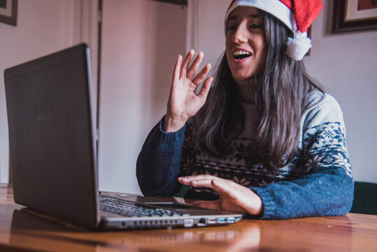 Una chica joven haciendo una video llamada saluda a su ordenador portátil mientras lleva puesto un gorro de Santa Claus y sujeta su teléfono móvil - Powered by Adobe