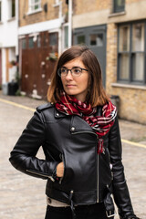 Obraz premium Portrait of an attractive young female wearing a black leather jacket, a red scarf and glasses in a small alley in London, United Kingdom