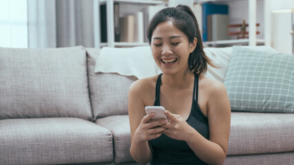 Happy strong athletic fitness woman in sport top using mobile phone after morning exercises at home in bright living room