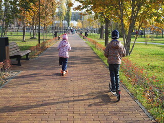Fototapeta premium Happy children ride a scooter in the park. A boy and a girl rest in the autumn outdoors. Back view.
