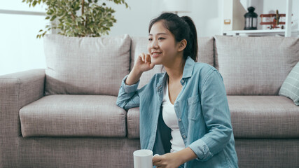 lovely smiling young asian chinese woman sitting by couch is enjoying carefree sunday with cup of hot coffee.