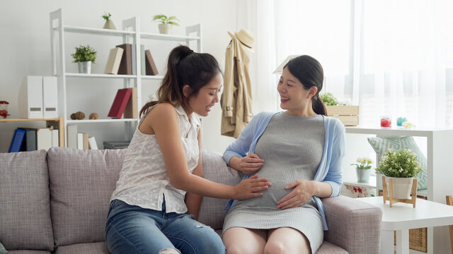 Beautiful pregnant woman and girl best friend smoothing tummy and having fun during baby shower at home living room.