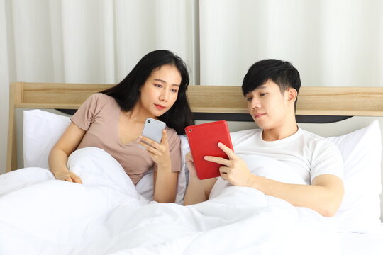 Asian couple looking at each other phone while laying down in the bed planning for their upcoming wedding 