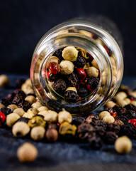 Close-up of 4 kinds of peppercorns spilling out of a glass bottle on a dark textured surface