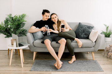 Smiling young Caucasian sporty couple sitting on gray sofa at cozy room at home and holding bottles of water, resting after workout, and using smartphone, watching video