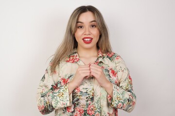 Dreamy charming young blonde woman standing against white background with pleasant expression, keeps hands crossed near face, excited about something pleasant.