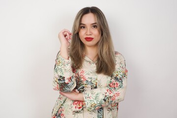 Image of cheerful pretty young woman standing against gray wall with hand near face. Looking with glad expression at the camera after listening to good news. Confident girl.