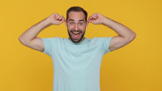 Fun young man in basic casual blue t-shirt isolated on yellow background studio. People emotions lifestyle concept. Looking camera fooling around pointing index fingers on blowing cheeks monkey ears