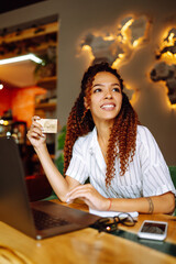 African American woman with credit card and laptop computer doing online shopping sitting at cafe or restaurant. Online shopping, e-commerce, internet banking, spending money.
