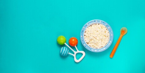 Bowl with cottage cheese, baby food, rattle and spoon, on a blue background, top view, horizontal, 