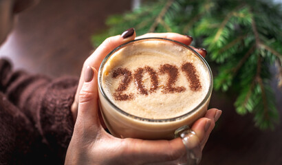 Women's hands are holding a transparent cup of coffee. There are numbers 2021 on the cinnamon milk...
