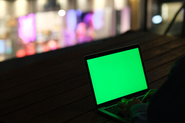 over shoulder of hands typing green screen laptop computer at dark night. Colorful city lights bokeh background