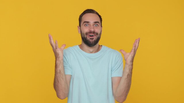Bearded happy young man 20s in basic casual blue t-shirt looking shocked surprised wow showing hands brain explosion head isolated on yellow background studio. People sincere emotion lifestyle concept