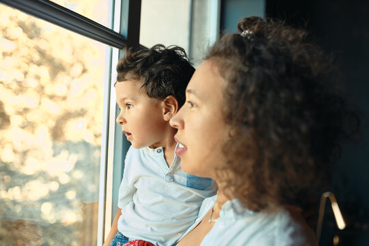Indoor image of happy young Latin mother staying home standing by window with preschool baby son in her arms, looking outside through glass. Motherhood, parenting, children and family concept