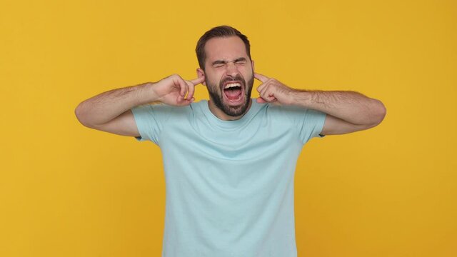 Sad mad crazy displeased young man 20s years old in basic blue t-shirt closed eyes cover ears do not want to listen screaming isolated on yellow background in studio. People emotions lifestyle concept