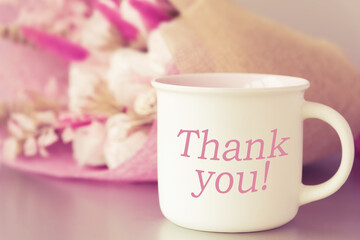 Fototapeta na wymiar Focus on cup with coffee or tea with thank you inscription. Mockup mug with space space for text. Blurry flower bouquet on table. Cafe or restaurant with tasty beverages and hot warming drinks