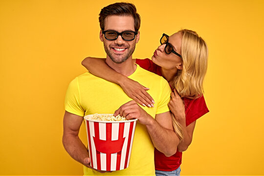 Laughing couple two friends guy girl in 3d glasses isolated on yellow background. People in cinema lifestyle concept. Watching movie film holding bucket of popcorn, hugging, kiss in cheek