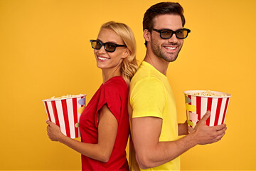 Smiling young couple two friends guy girl in 3d glasses posing isolated on yellow background. People in cinema lifestyle concept. Watching movie film, hold buckets of popcorn standing back to back