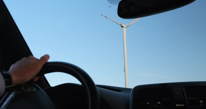 A man drives an electric car into the area of wind farms operation. Video shooting from behind the driver's shoulder inside the car.
