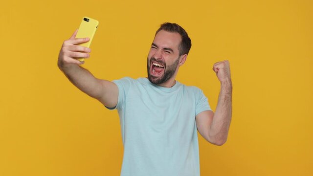 Fun confident charismatic happy bearded young man in basic casual blue t-shirt posing doing selfie shot on mobile cell phone video call isolated on yellow background studio. People lifestyle concept