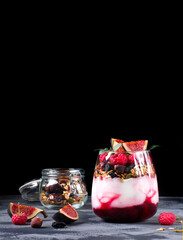 Layered trifle dessert in the glass on the grey table. Cherry jam, natural yogurt, granola and raspberry topped with figs and mint. Low key 