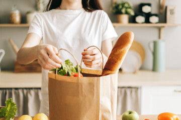 Caucasian woman hold eco shopping bag with fresh vegetables and baguette in modern kitchen.