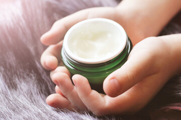 Baby boy little hands holding white cream bottle on grey fur background.