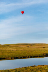 Hot air balloon in the shape of a heart is landing behind the lake, mirroring in it