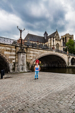 Latin woman with the St Michael bridge with its arches and the Leie river in the background, visiting the city of Ghent, Belgium on a cloudy day with a gray sky with heavy clouds