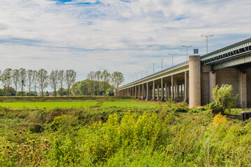 Fototapeta na wymiar Road bridge A76 (Scharbergbrug) over the Maas river field with green grass and trees, sunny day with blue sky and abundant white clouds in Elsloo and Meers, South-Limburg in the Netherlands Holland