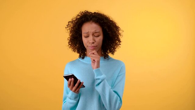 puzzled young woman on a yellow background touches her chin and looks into a smartphone. The woman makes a choice between several plaques. Isolated on a bright yellow background. 4K