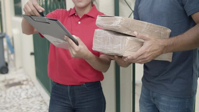 Female delivery worker in red uniform bringing package to customers door. Man opening gate, taking parcels and signing for receiving. Shipping or delivery service concept