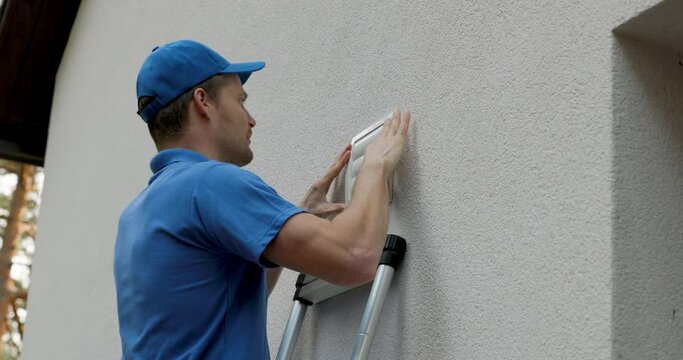 home ventilation - hvac worker standing on ladder and installing air duct grille on house facade