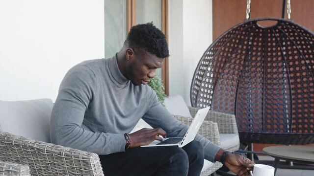 Young handsom black man working on the laptop on his balcony while drinking coffee. High quality 4k footage