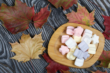 Marshmallows of different colors. They lie on black pine boards. Autumn maple leaves are scattered around.