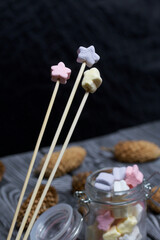 Marshmallows of different colors. They lie in a glass jar. One is strung on a wooden skewer. Nearby are spruce cones.