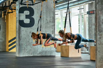 Two young athletic girls exercising with trx fitness straps at gym