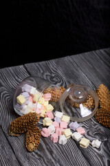 Marshmallows of different colors. They lie in an overturned glass jar. Nearby are spruce cones. Against the background of black pine boards.