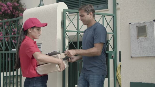Positive courier girl in red delivering package to customers door. Man opening gate, taking parcels and signing for receiving. Shipping or delivery service concept