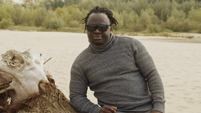 Attractive african man with sunglasses resting on the sandy beach in autumn. Leaning on the old dry tree trunk